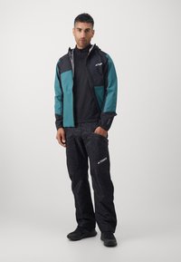 Ανδρική εξωτερική ένδυση: πολύχρωμο μπουφάν με τιρκουάζ και μαύρες πάνελ, συνδυασμένο με μοτίβο μαύρα παντελόνια. Διαθέτει ανθεκτικό ύφασμα και ανακλαστικές λεπτομέρειες.