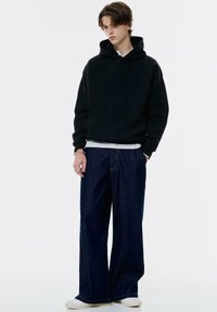 Giovane uomo che indossa un hoodie nero, jeans larghi blu scuro, sneakers bianche e un braccialetto d'argento, in piedi contro uno sfondo bianco uniforme.