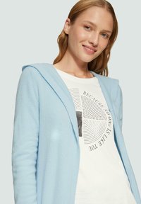Hellblauer Kapuzenpulli aus weichem Material, mit geribbelter Textur und offenem Front, darüber getragen ein weißes T-Shirt mit schwarzem Druck.