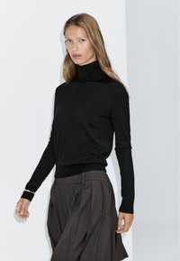Maglione a collo alto nero in morbido materiale lavorato a maglia, con maniche lunghe e silhouette aderente, abbinato a pantaloni grigi scuri con pieghe.