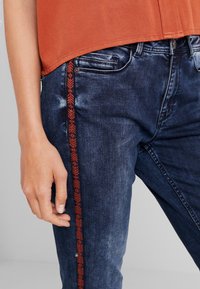 Los jeans de mezclilla azul oscuro presentan un patrón bordado en rojo a lo largo de la costura lateral. Una blusa suelta de color óxido cubre parcialmente la cinturilla.