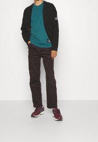 Cardigan nero sopra una maglietta teal, abbinato a pantaloni in corduroy scuri e scarpe da corsa bordeaux con dettagli bianchi e riflettenti.