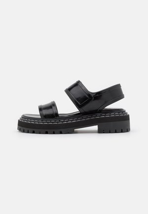 Sort sandal med chunky sål med to brede remme og en rem bagpå, med hvide syninger langs sålens kant, vist fra siden.