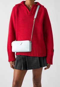 Červený pletený svetr s výstřihem do V a žebrovaným lemem, doplněný černou kladovanou plisovanou sukní. Malá světle modrá kabelka s řetízkovým popruhem je nošena přes tělo.