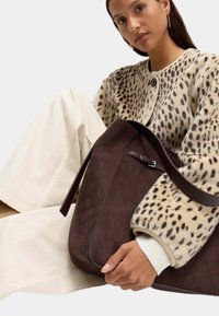 Hnědá semišová taška s hnědými koženými detaily, se strukturovaným tvarem a širokým ramenním popruhem, držena proti svetr s leopardím vzorem.