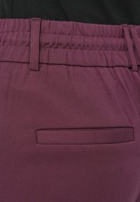 Pantaloni color marrone scuro con vita elasticizzata, caratterizzati da una tasca posteriore piatta e una texture liscia. Tessuto liscio con design aderente.