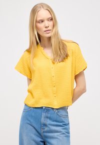 Gul, kortærmet top med en struktureret stof, knapfront og en cropped pasform, parret med lyseblå jeans.