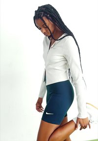 Balta šiurkšti zip-up striukė derinama su tamsiai mėlynomis dviratininko šortais, su baltu Nike logotipu. Modelis tempia vieną koją.