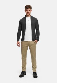 Šedý svetr na zapínání na zip s pleteným vzorem, nošený přes bílé tričko. Kombinován s béžovými chino kalhotami a černými botami, stojící před neutrálním pozadím.