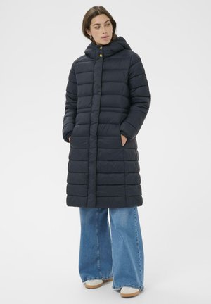 Langer Daunenmantel in Dunkelblau mit hohem Kragen, horizontalen Steppnähten und Seitentaschen. Kombiniert mit ausgestellten blauen Jeans und beigen Schuhen.
