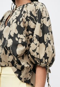 Luźna bluzka w kwiatowy wzór w beżowo-czarnych kolorach. Posiada bufiaste rękawy, dekolt w kształcie litery V oraz wiązania na mankietach. Miękka tekstura tkaniny.