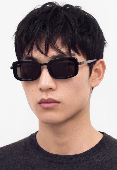 Jeune homme aux cheveux courts noirs portant des lunettes de soleil rectangulaires noires et un t-shirt sombre à col rond sur un fond clair uni.
