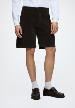 Homme portant un short noir longueur genoux, des chaussettes blanches montantes, des chaussures en cuir marron et une chemise blanche à manches longues sur un fond uni.