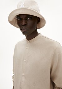 Beige katoenen overhemd met knopen en staande kraag, voorzien van een geborduurd logo, gecombineerd met een bijpassende bucket hat, beide met een gladde textuur.