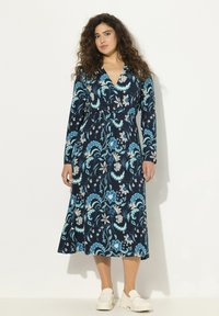 Granatowa sukienka w kwiaty z długimi rękawami, dekoltem w kształcie litery V oraz zwiewnym krojem z niebieskimi i złotymi akcentami. Białe buty wsuwane.