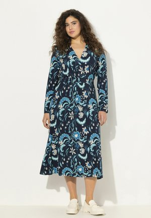 Robe portefeuille florale marine avec manches longues, décolleté en V et silhouette fluide agrémentée de accents bleus et dorés. Chaussures blanches à enfiler.
