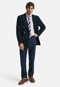 Marineblå stribet dobbeltbrystet blazer, stribet skjorte og slips, parret med mørke bukser og brune sko. Gyldne knapper på blazeren.