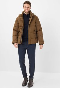 Veste rembourrée marron avec un col haut, texture matelassée et accents noirs, associée à un pull navy et un pantalon navy, complétée par des bottes marron.