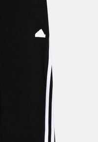 Pantaloni sportivi neri con strisce laterali bianche e una toppa con logo bianco. Il tessuto appare morbido e liscio con un design aderente.