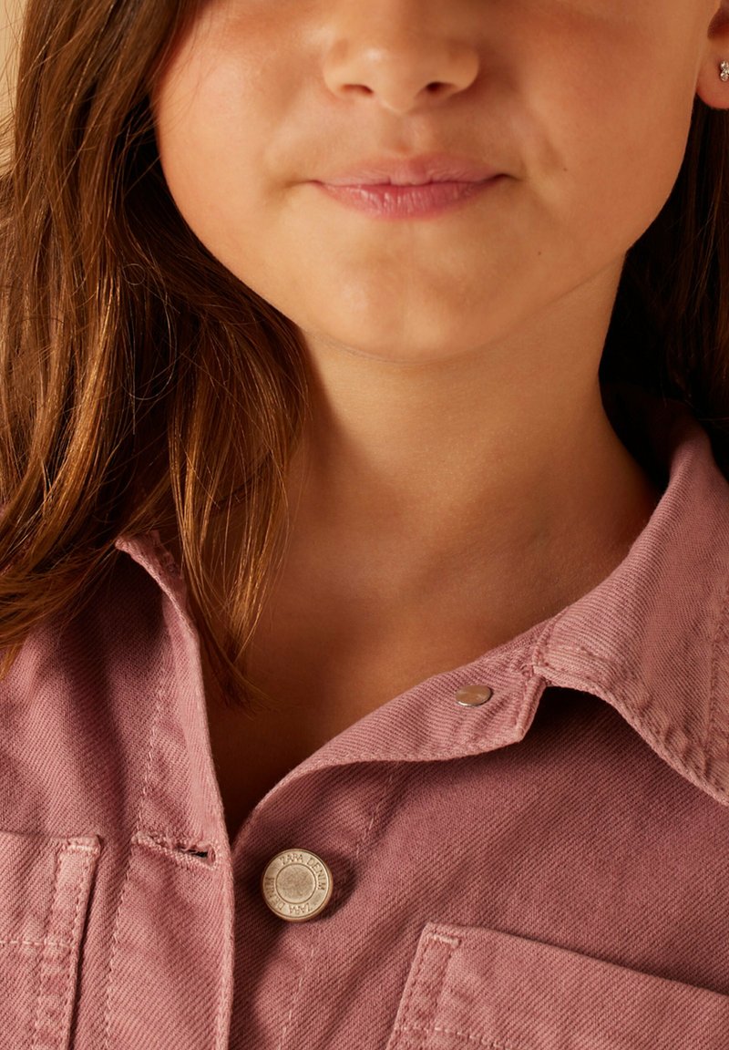 Lichtroze corduroy shirt met een kraag, voorzien van een zilveren knoop, subtiele textuur en een detail van een borstzak. Close-up van de halslijn.