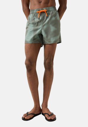 Mandlig model iført grønne badeshorts med orange snor og sorte klipklappere, vist fra midten af torsoen til fødderne på ensfarvet baggrund.