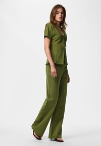 Jeune femme portant un chemisier olive à manches courtes boutonné et un pantalon large assorti avec des sandales à talons noires.
