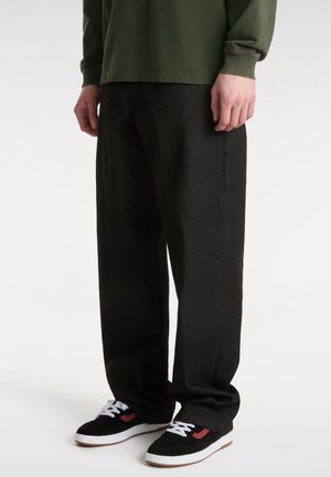 Persona che indossa pantaloni neri larghi, sneakers nere con suole e lacci bianchi e una maglia verde scuro a maniche lunghe che sta in piedi su un pavimento bianco.
