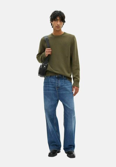 Joven vestido con un suéter desgastado de color verde olivo, jeans azules holgados, zapatos negros y una bolsa de hombro negra sobre un fondo blanco.