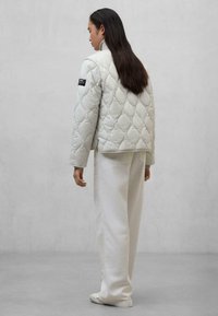 Lichtgrijze gewatteerde jacket met ronde vormen, hoge kraag en zwart label op de linker mouw, gerapporteerd met wijde witte broeken.