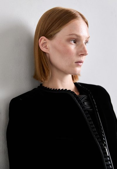 Chaqueta de terciopelo negro con detalle de cuello con volantes y acentos brillantes, llevada por un modelo con cabello rubio rojizo hasta los hombros frente a un fondo neutro.
