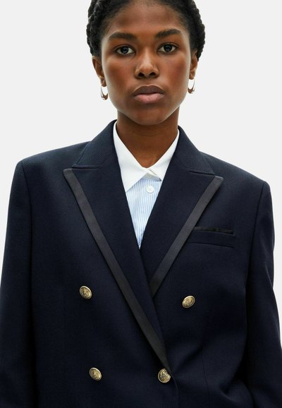 Blazer navy a doppio petto con revers in raso, bottoni dorati e taschino sul petto. Camicia bianca con colletto e righe blu sotto.