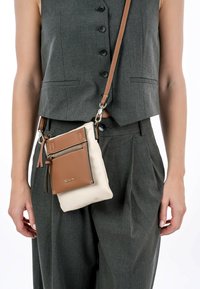 Rintbag i beige og brun læder med en frontlomme med lynlås, frynser detalje og justerbar rem, båret mod et gråt sæt tøj.