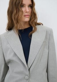 Lichtgrijze blazer met een gestructureerd patroon, uitgesproken schoudernaden en een grote knop, gedragen over een donkerblauwe trui.