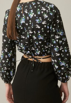 Femme portant un crop top noir à manches longues à fleurs, noué dans le dos, et une jupe noire taille haute, vue de dos.