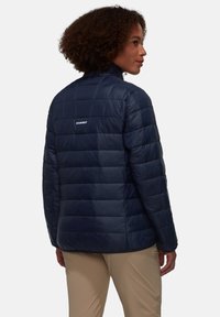 Giacca puffer blu navale isolata con design trapuntato, colletto alto e logo bianco sul retro. Tessuto sintetico liscio con una leggera lucentezza.