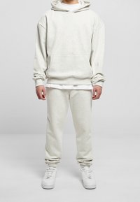 Uomo che indossa un hoodie grigio chiaro e pantaloni jogger abbinati con sneakers bianche, in piedi su uno sfondo grigio uniforme.