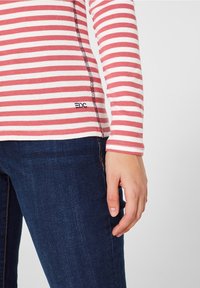 Camicia a maniche lunghe a righe rosa e bianche con una cucitura nera e un piccolo dettaglio del logo. Abbinata a jeans in denim blu scuro.