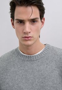 Suéter de punto gris con un cuello acanalado. Textura suave, corte holgado, superpuesto a una camisa blanca. Líneas limpias y diseño minimalista.