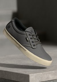 Sapatilha de tela cinza com bico redondo, sola de borracha bege, atacadores na parte da frente e detalhe do logotipo na lateral. Superfície texturizada e acabamentos cosidos.