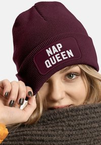 Maroon knit beanie with a wide, rectangular patch displaying "NAP QUEEN" in bold white text. Soft texture with fitted design.