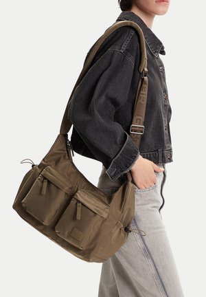 Person trägt eine schwarze Jeansjacke und helle Jeans und trägt eine große olivgrüne Umhängetasche mit zwei vorderen Reißverschlusstaschen und verstellbarem Riemen.