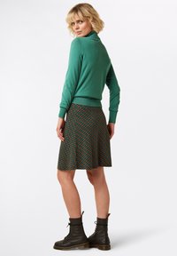 Grüner, taillierter Rollkragenpullover kombiniert mit einem gemusterten Rock in Dunkelgrün, Orange und Blau. Das Model trägt schwarze Schnürstiefel.