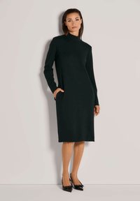 Schwarzes, langärmeliges Kleid mit hohem Kragen und seitlichen Taschen. Glatte Textur, knieumspielend, getragen mit schwarzen, spitzen Absatzschuhen.