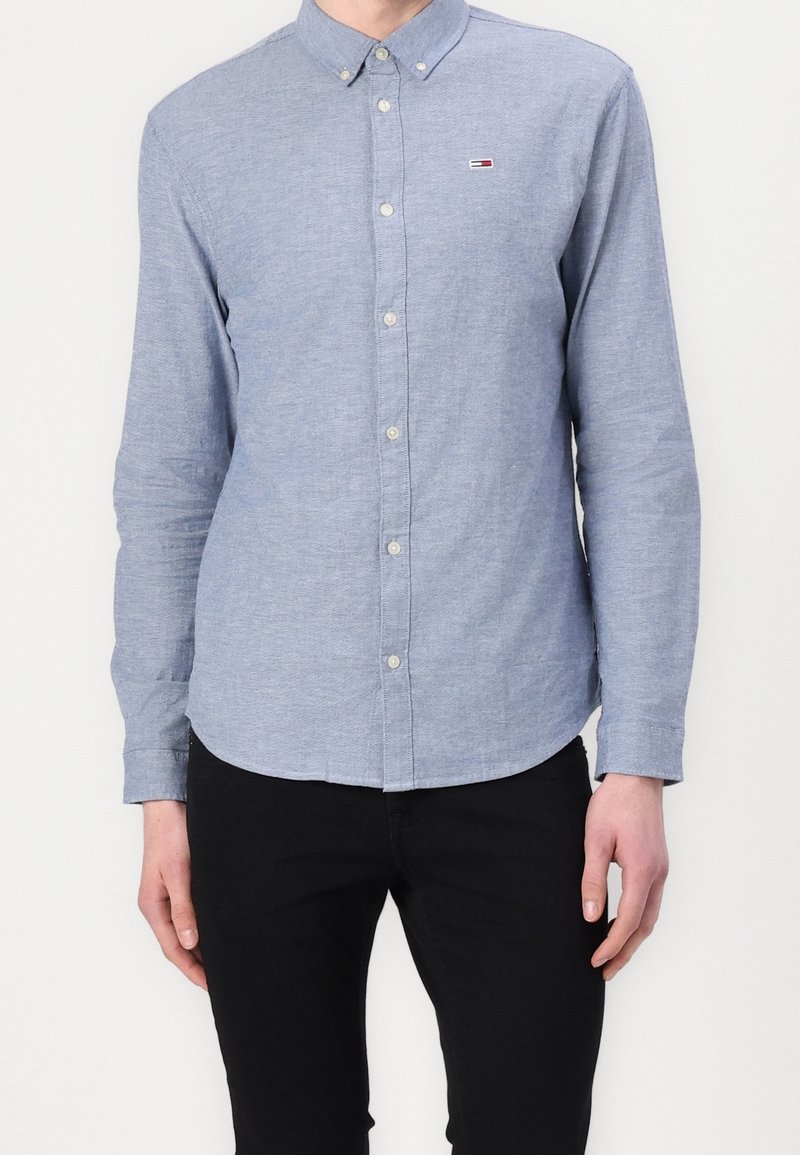 Camisa de botones azul claro hecha de un tejido suave, con un collar clásico y un pequeño parche con el logo en el lado izquierdo del pecho. Mangas largas.