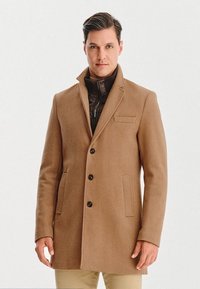 Karmelowy płaszcz z wełny z wykładanym kołnierzem, pi ęcioma czarnymi guzikami i bocznymi kieszeniami. Nałożony na ciemną kurtkę z zamkiem błyskawicznym.
