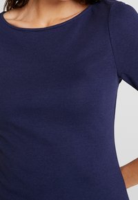 Marinblå, figurnära topp i mjukt, stretchigt material. Har rund halsringning och långa ärmar, med en slät textur och minimala detaljer.