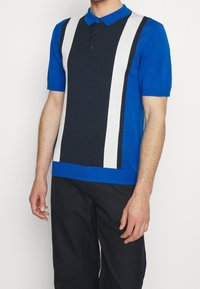 Modrá polo košile s krátkým rukávem, zdobená námořnicky modrými a bílými vertikálními pruhy. Vyrobena z texturovaného materiálu s límečkem a třemi knoflíky.