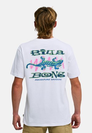 Person mit kurzen braunen Haaren, die ein weißes T-Shirt trägt, das eine blaue und grüne Eidechsen-Grafik und den Schriftzug "Billabong Adventure Division" auf der Rückseite zeigt.