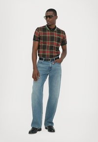 Cămașă polo cu carouri negre și roșii, cu mâneci scurte, asortată cu blugi drepți de culoare albastră deschisă și pantofi negri, accesorizată cu ochelari de soare.