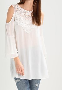 Blouse blanche avec des détails en dentelle au niveau du cou et des épaules, manches longues en forme de cloche et texture légère. Associée à un jean bleu déchiré.